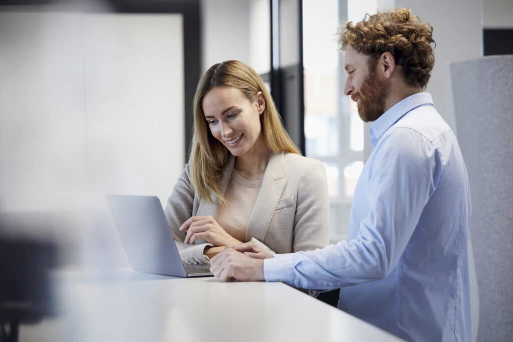 Two smiling business people are looking something up in their company's intranet at a laptop in an office.