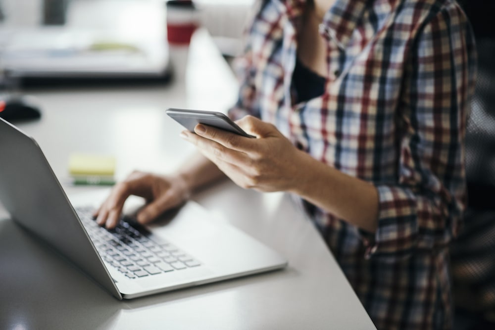 Junge Frau arbeit an ihrem Computer und schaut sich etwas auf ihrem Smartphone an.