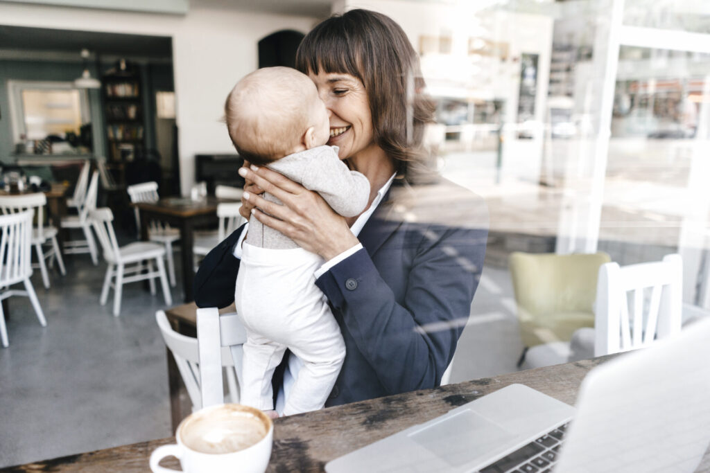 Lächelnde Geschäftsfrau sitzt in einem Café mit ihrem Baby auf dem Arm.