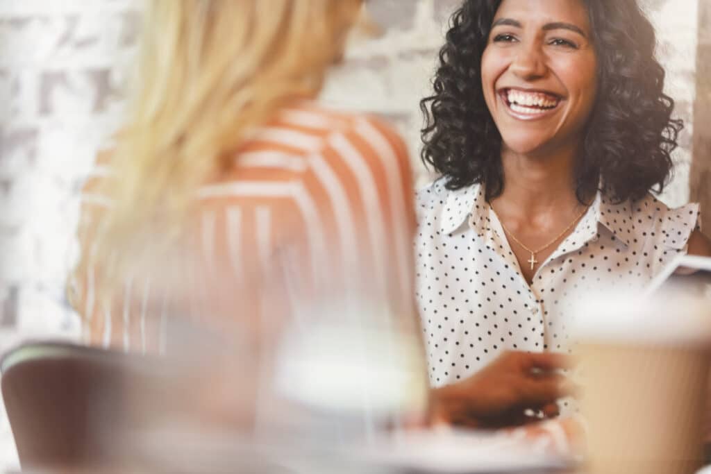 Zwei lächelnde Mitarbeiterinnen sitzen in einem Café und freuen sich über ihre Workation.