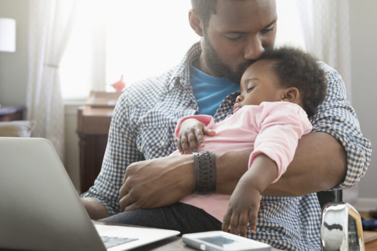 A father sitting at a computer, holding his baby and kissing its forehead.