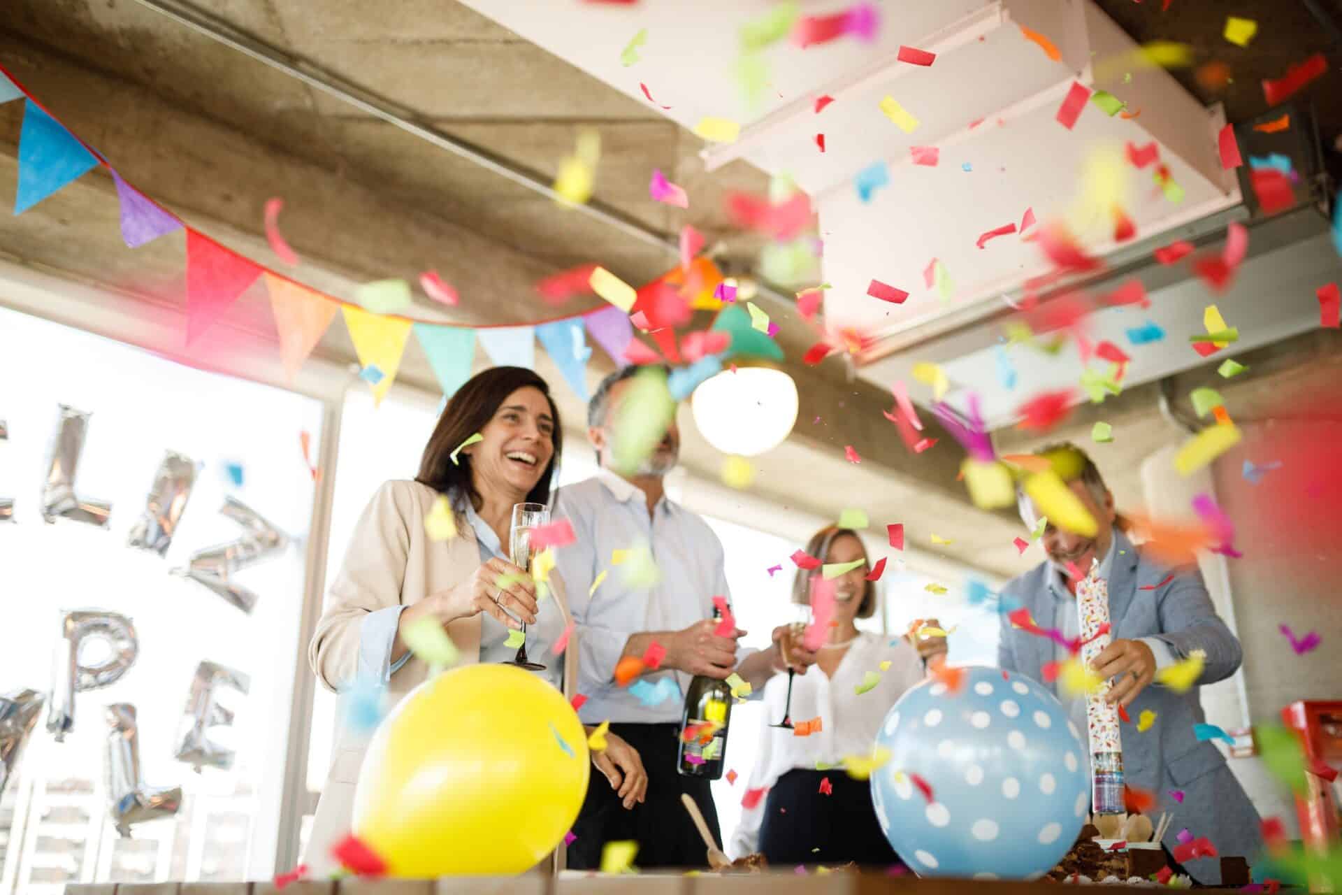 Fröhliche Mitarbeiter:innen feiern im Büro ein Jubiläum mit Konfetti und Geschenken.