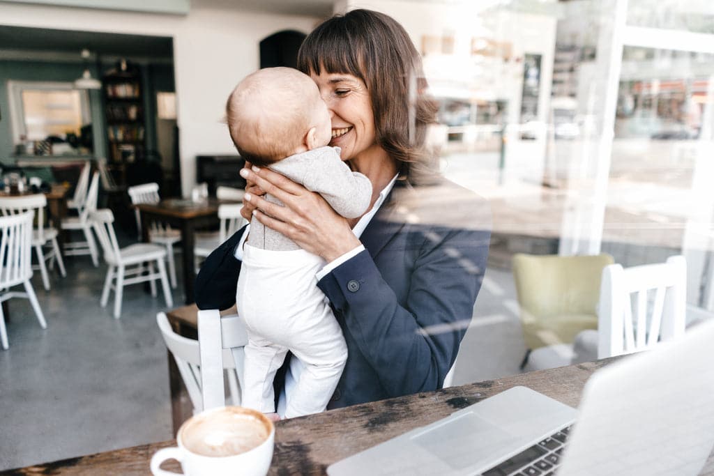 Glückliche Geschäftsfrau sitzt am Computer und hält ihr Baby hoch.