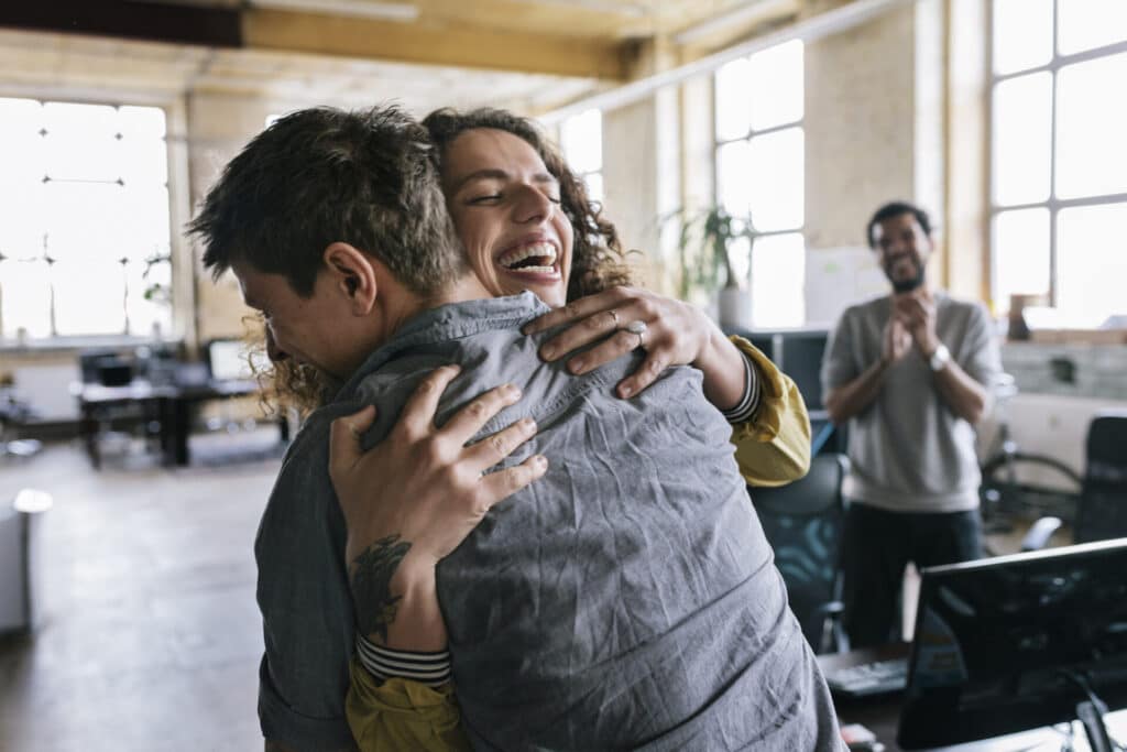 Smiling colleagues are hugging in an office.