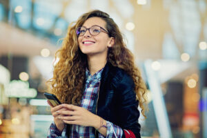Young adult woman is smiling and holding her smartphone, as she just had a great customer experience.