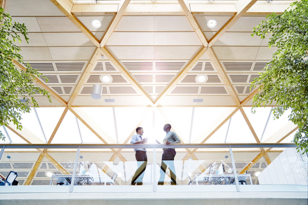 Zwei Arbeitskollegen stehen in einem modernen Büro mit Holzpaneelen und großen grünen Pflanzen.
