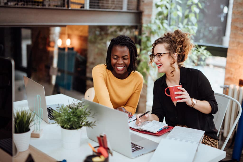 Zwei junge, lächelnde Geschäftsfrauen unterhalten sich an ihrem Arbeitsplatz im Büro.